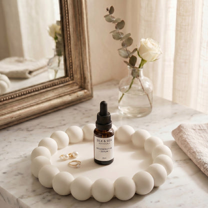 Skincare dropper bottle on a decorative white tray with a mirror and flowers in the background.